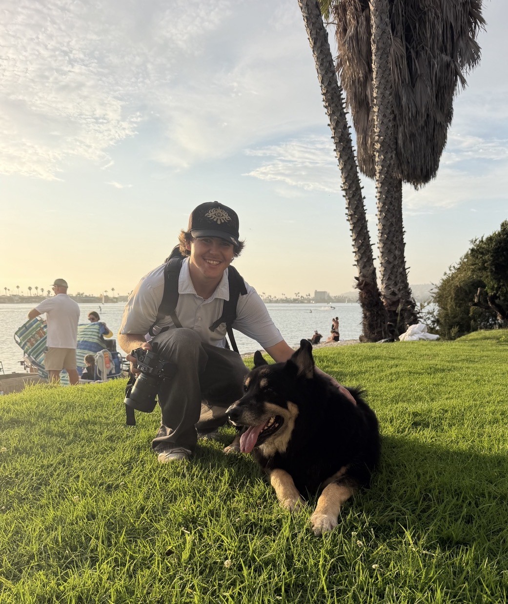 Zach Jenkins kneeling on the grass with a dog by the water, camera in hand
